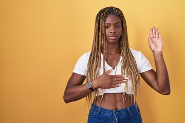 African american woman with braided hair standing over yellow background swearing with hand on...