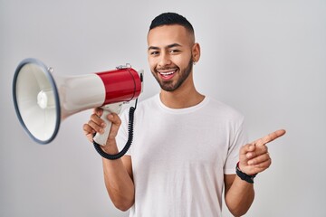 Young hispanic man shouting through megaphone smiling happy pointing with hand and finger to the side