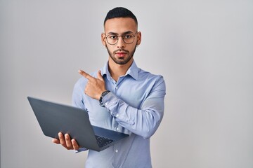 Young hispanic man working using computer laptop pointing with hand finger to the side showing advertisement, serious and calm face