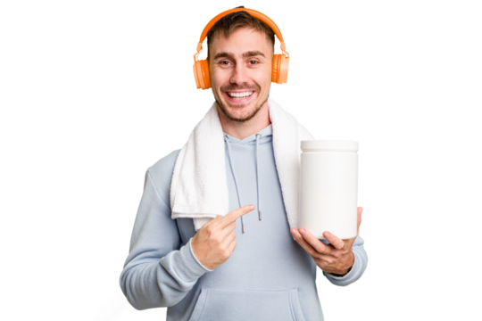 Young fitness man listening to music and holding a protein bottle isolated cut out smiling and pointing aside, showing something at blank space.