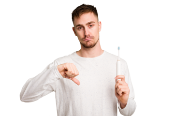 Young caucasian man holding an electric toothbrush isolated cut out showing a dislike gesture, thumbs down. Disagreement concept.