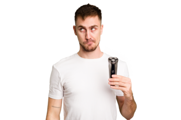 Young man holding a shaving machine cut out isolated confused, feels doubtful and unsure.