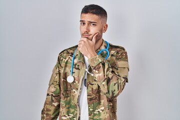 Young hispanic doctor wearing camouflage army uniform thinking concentrated about doubt with finger...
