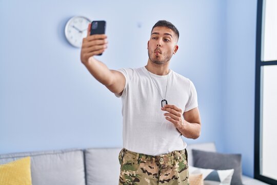Young hispanic man wearing camouflage army uniform taking selfie at home making fish face with mouth and squinting eyes, crazy and comical.