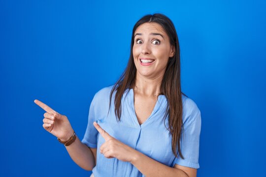 Young brunette woman standing over blue background pointing aside worried and nervous with both hands, concerned and surprised expression