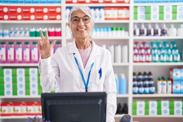 Middle age woman with tattoos working at pharmacy drugstore showing and pointing up with fingers number three while smiling confident and happy.
