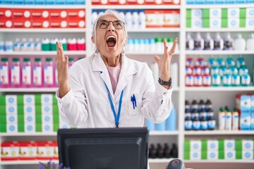 Middle age woman with tattoos working at pharmacy drugstore crazy and mad shouting and yelling with aggressive expression and arms raised. frustration concept.