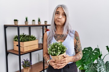 Middle age grey-haired woman holding green plant pot at home puffing cheeks with funny face. mouth inflated with air, crazy expression. - obrazy, fototapety, plakaty