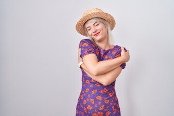 Young caucasian woman wearing flowers dress and summer hat hugging oneself happy and positive,...