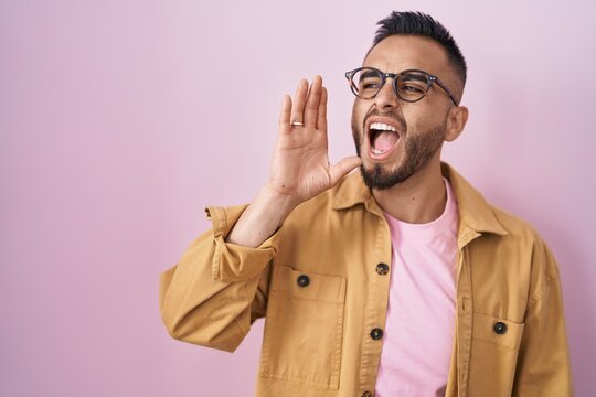 Young hispanic man standing over pink background shouting and screaming loud to side with hand on mouth. communication concept.