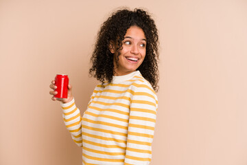 Young african american woman holding a cola refreshment isolated looks aside smiling, cheerful and pleasant. - obrazy, fototapety, plakaty