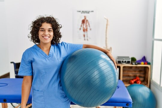 Young hispanic woman wearing physio therapist uniform holding fit ball at clinic