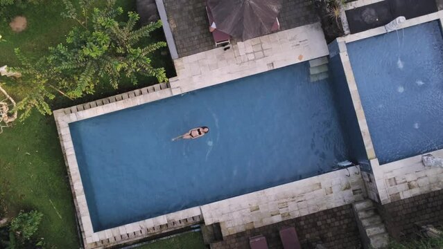 Girl in black bikini is swimming in blue pool among jungles in Bali, Indonesia. Aerial view, drone shot, zoom in
