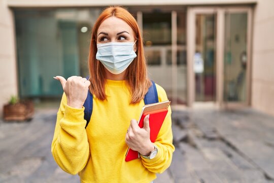 Young woman wearing safety mask and student backpack holding books pointing thumb up to the side smiling happy with open mouth