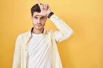 Young hispanic man standing over yellow background making fun of people with fingers on forehead...