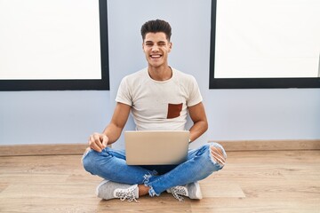Young hispanic man using laptop at home winking looking at the camera with sexy expression,...