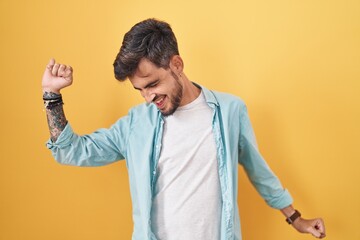 Young hispanic man with tattoos standing over yellow background dancing happy and cheerful, smiling...