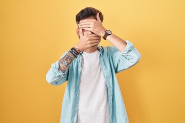 Young hispanic man with tattoos standing over yellow background covering eyes and mouth with hands, surprised and shocked. hiding emotion