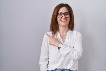 Brunette woman standing over white isolated background cheerful with a smile on face pointing with hand and finger up to the side with happy and natural expression