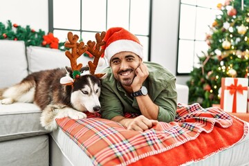 Obraz premium Young hispanic man lying on sofa with dog wearing reindeer hat by christmas tree at home