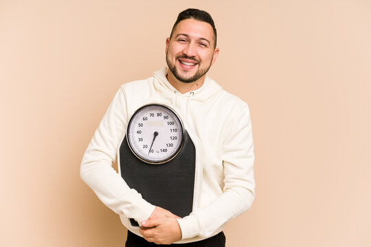 Adult latin man holding a weight scale isolated laughing and having fun.