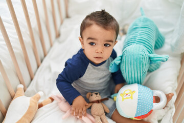 Adorable hispanic toddler sitting on cradle with relaxed expression at bedroom