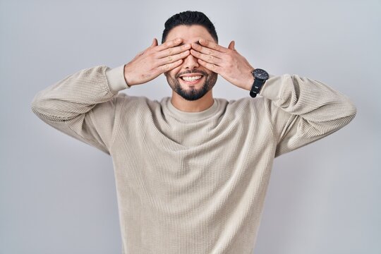 Young handsome man standing over isolated background covering eyes with hands smiling cheerful and funny. blind concept.