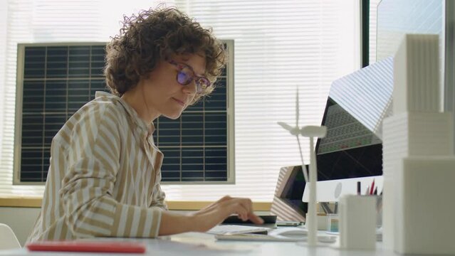 Female renewable energy developer typing on computer and using digital tablet while working in office with solar batteries
