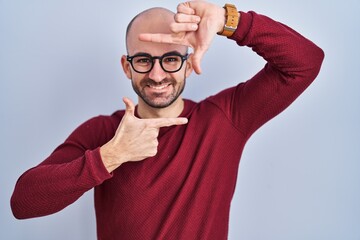 Young bald man with beard standing over white background wearing glasses smiling making frame with hands and fingers with happy face. creativity and photography concept.