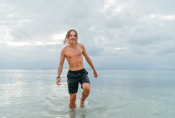 Naklejka premium Smiling muscular longhaired teenage boy walking out from Indian ocean waves after evening swimming. Traveling and an exotic vacations concept image.