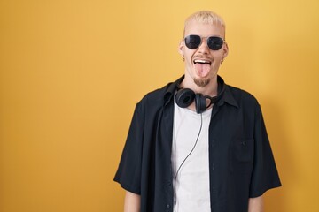 Young caucasian man wearing sunglasses standing over yellow background sticking tongue out happy with funny expression. emotion concept.