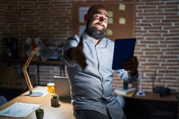 Young hispanic man with beard and tattoos working at the office at night smiling friendly offering handshake as greeting and welcoming. successful business.