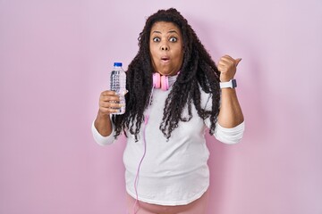 Plus size hispanic woman wearing sportswear and headphones surprised pointing with hand finger to the side, open mouth amazed expression.