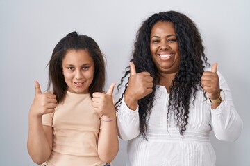 Mother and young daughter standing over white background success sign doing positive gesture with hand, thumbs up smiling and happy. cheerful expression and winner gesture.