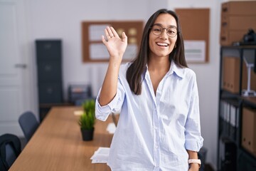 Young hispanic woman at the office waiving saying hello happy and smiling, friendly welcome gesture - obrazy, fototapety, plakaty