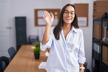 Foto op Plexiglas Young hispanic woman at the office waiving saying hello happy and smiling, friendly welcome gesture © Krakenimages.com
