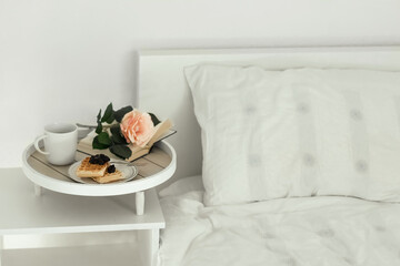 The atmosphere of a romantic morning. A cup of coffee, waffles, an open book and a flower on a wooden tray on a white bedside table near the bed. Domestic interior, hotel interior or medical study. Li