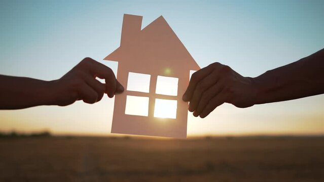 Happy family. People hands hold a paper house together. House made of paper is symbol of family happiness, mortgage protection, ecology. Sun shines through the window of house in nature at sunset
