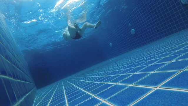 Male young adult tourist is floating on the surface of the water lying on the back in swimming pool filmed from the bottom in slow motion. View on caucasian man paddling with legs and arms from below