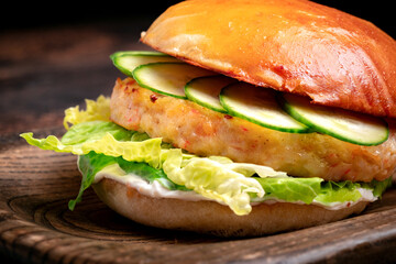 Burger with fish on a wooden board on a dark background. Bun with fish cutlet, cucumbers and mayonnaise, close up