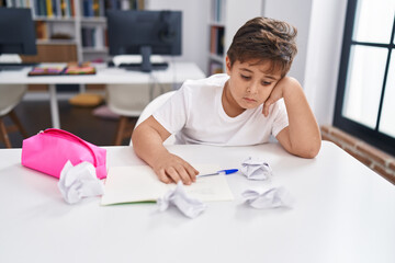 Adorable hispanic boy student sitting on table with worried expression at classroom