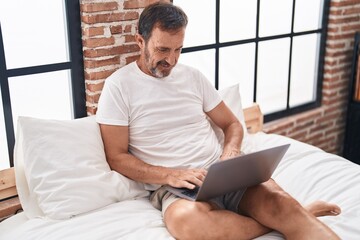 Middle age man using laptop sitting on bed at bedroom