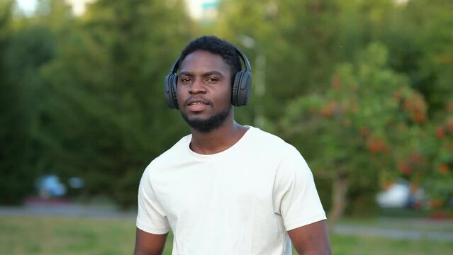 African American man wearing black headphones listens to favourite music. Bearded guy dances to excited music standing in green park