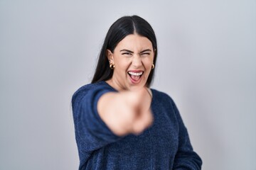 Young brunette woman standing over isolated background laughing at you, pointing finger to the camera with hand over body, shame expression