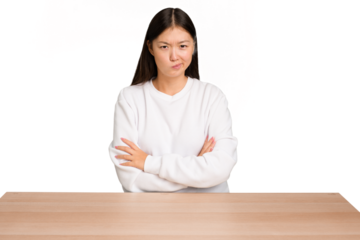 Young asian woman sitting on a table isolated frowning face in displeasure, keeps arms folded.