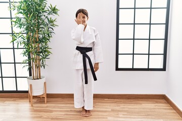 Fototapeta na wymiar Young hispanic girl wearing karate kimono and black belt covering one eye with hand, confident smile on face and surprise emotion.