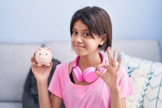 Young girl holding piggy bank doing ok sign with fingers, smiling friendly gesturing excellent symbol