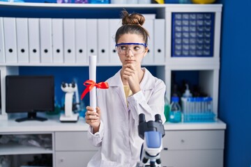 Teenager girl working at scientist laboratory holding degree serious face thinking about question with hand on chin, thoughtful about confusing idea