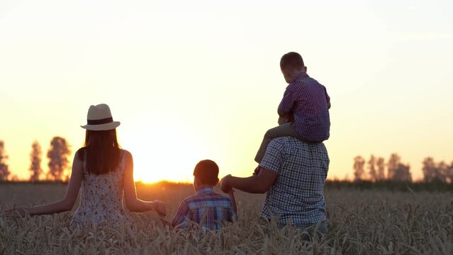 Parents silhouettes holding hands walk with lovely sons on wheat field at back sunset. Happy family explores countryside field and enjoys company