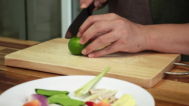Man is cutting juicy green lime with metal knife on kitchen chopping board filmed in closeup. Caucasian male hands are cooking meal carving sour fruit into pieces. Adding tropical products to dishes
